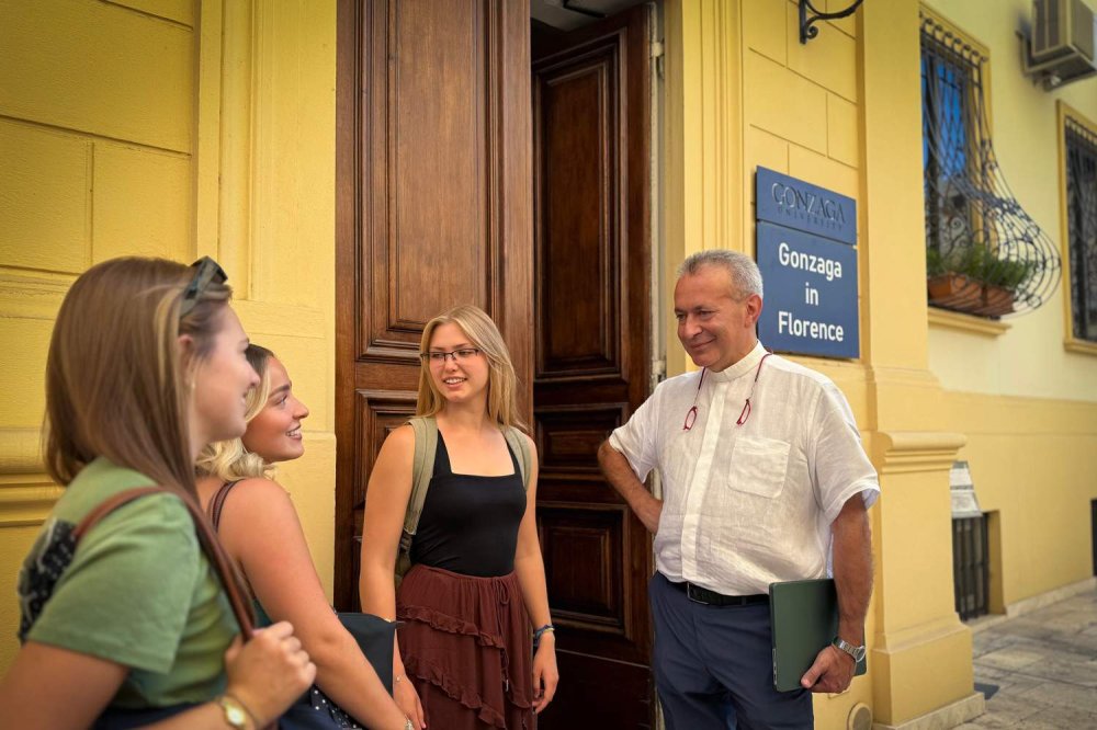 Don Alessandro Andreini sorridente con alcune studentesse davanti all'ingresso della Gonzaga University di Firenze