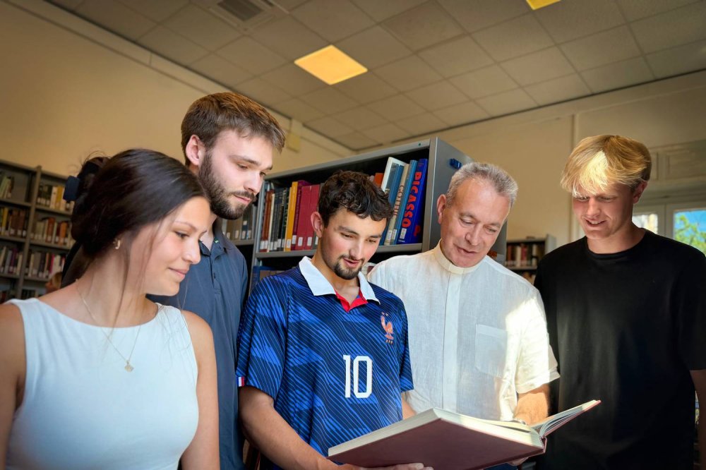 Don Alessandro Andreini sfoglia un libro con quattro studenti in una biblioteca